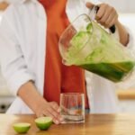 Close-up of a person pouring a vibrant green smoothie into a glass with limes on the wooden table.